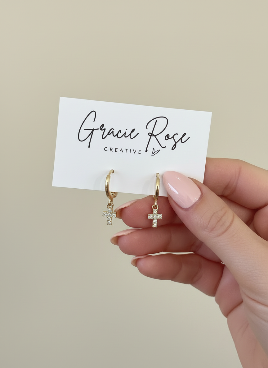 Hand holding Divine Huggies by Gracie Rose Creative, featuring gold huggie earrings with a cross charm, displayed against a neutral background.