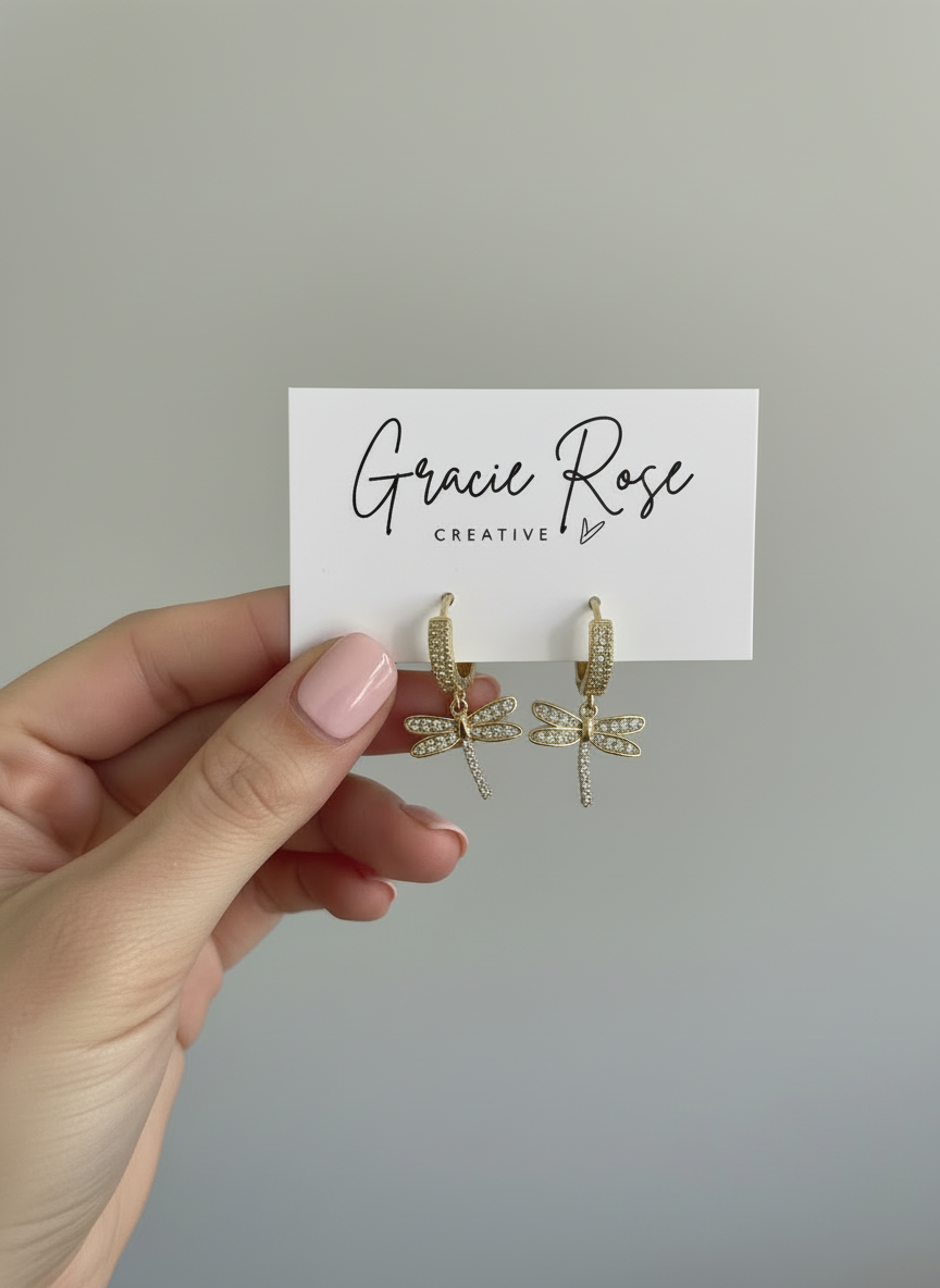 Hand holding Dragonfly Drops earrings by Gracie Rose Creative, displayed on a branded card, highlighting the intricate design of gold huggies and dragonfly charms.
