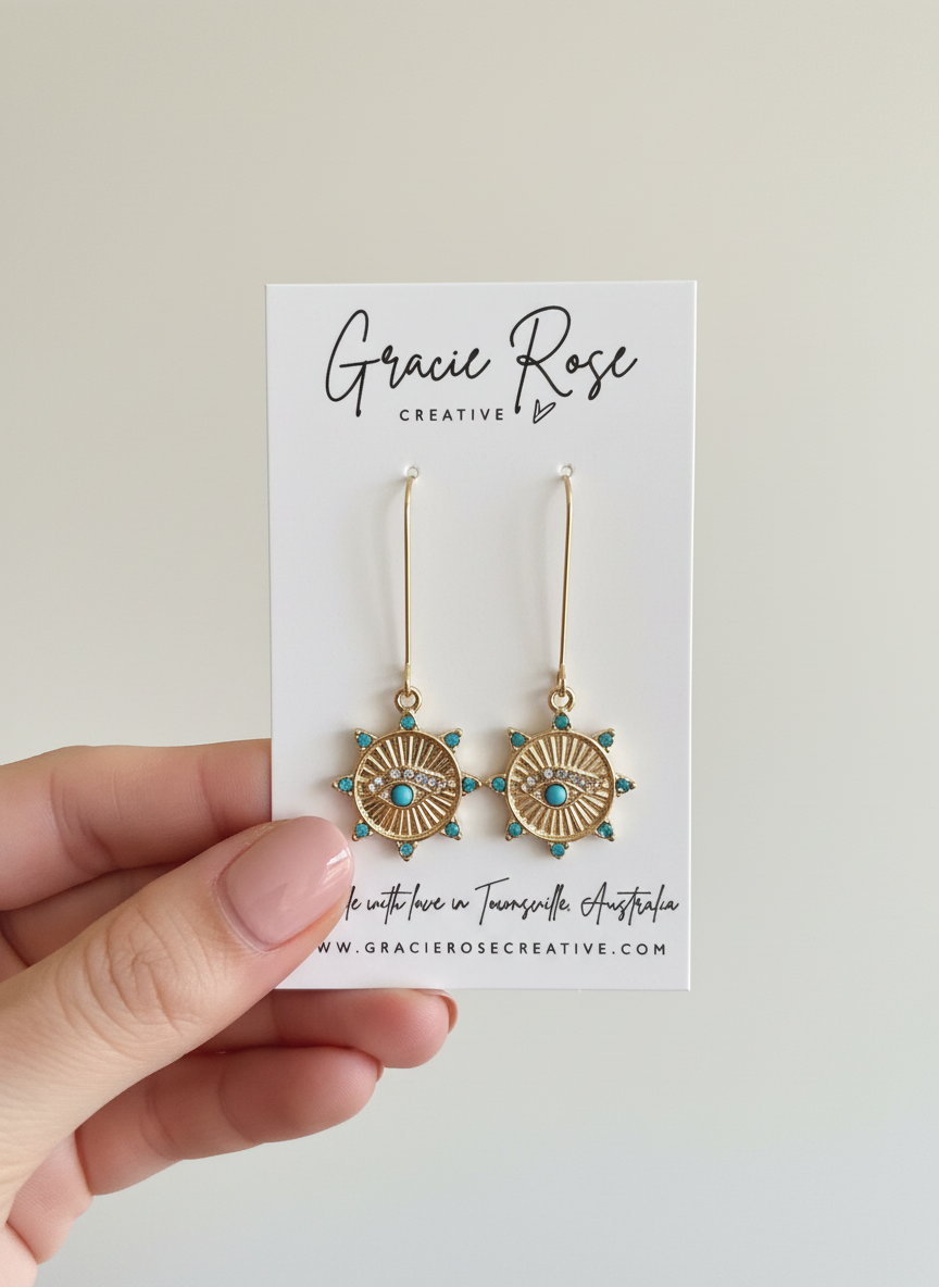 Handmade Solar Guardian Drops earrings by Gracie Rose Creative displayed on a branded card, showcasing the gold-toned sunburst and evil eye design.