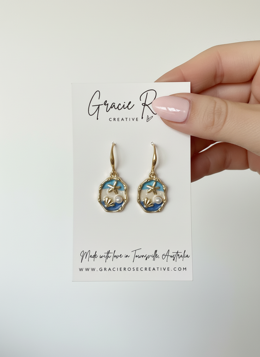 The Blue Horizon earrings by Gracie Rose Creative displayed on a hand, showing off the elegant seaside design with shell and pearl details.