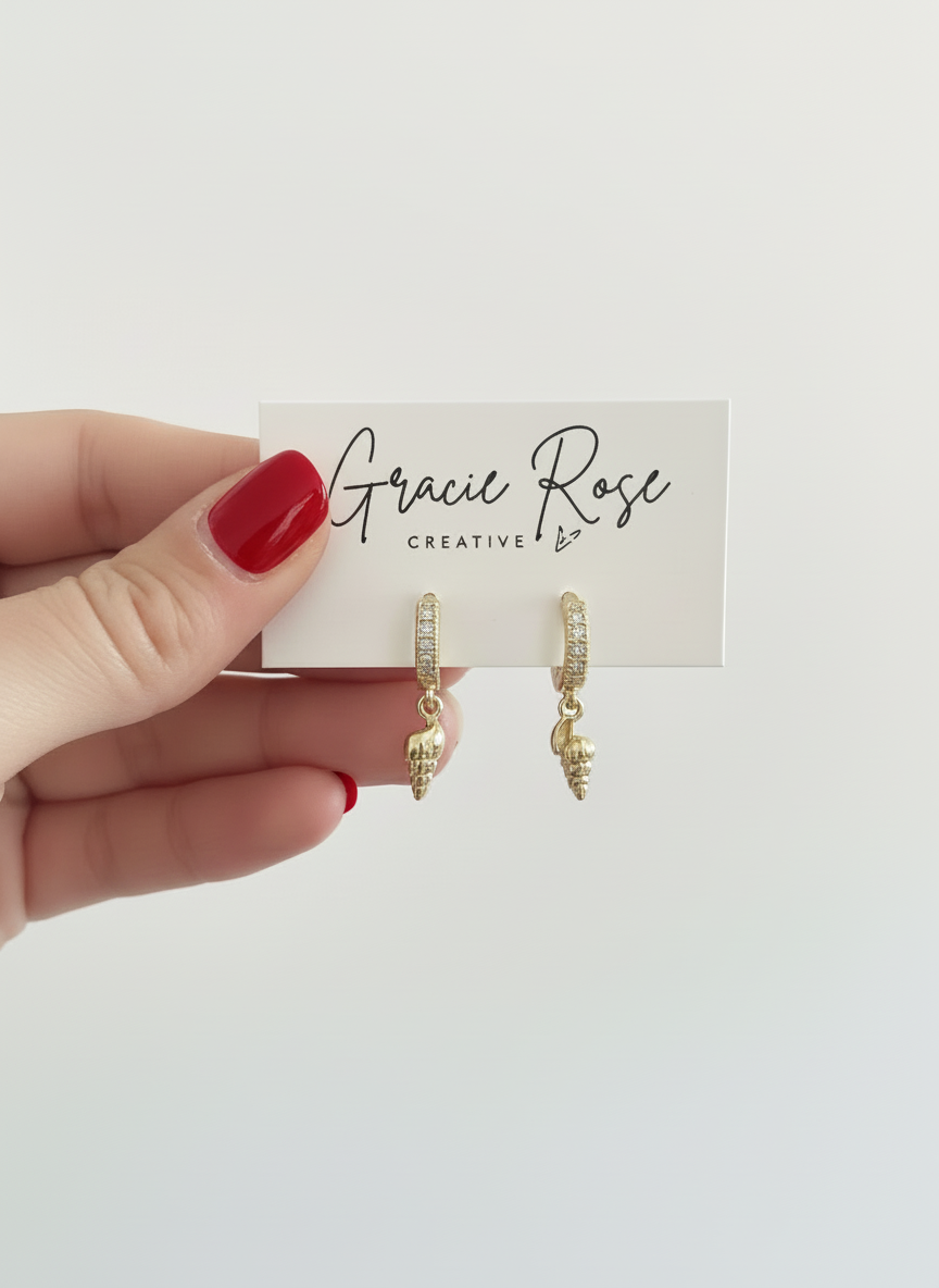 Hand holding Beach Babe Huggies earrings by Gracie Rose Creative against a white background, showcasing their elegant gold design and shell drop accents.
