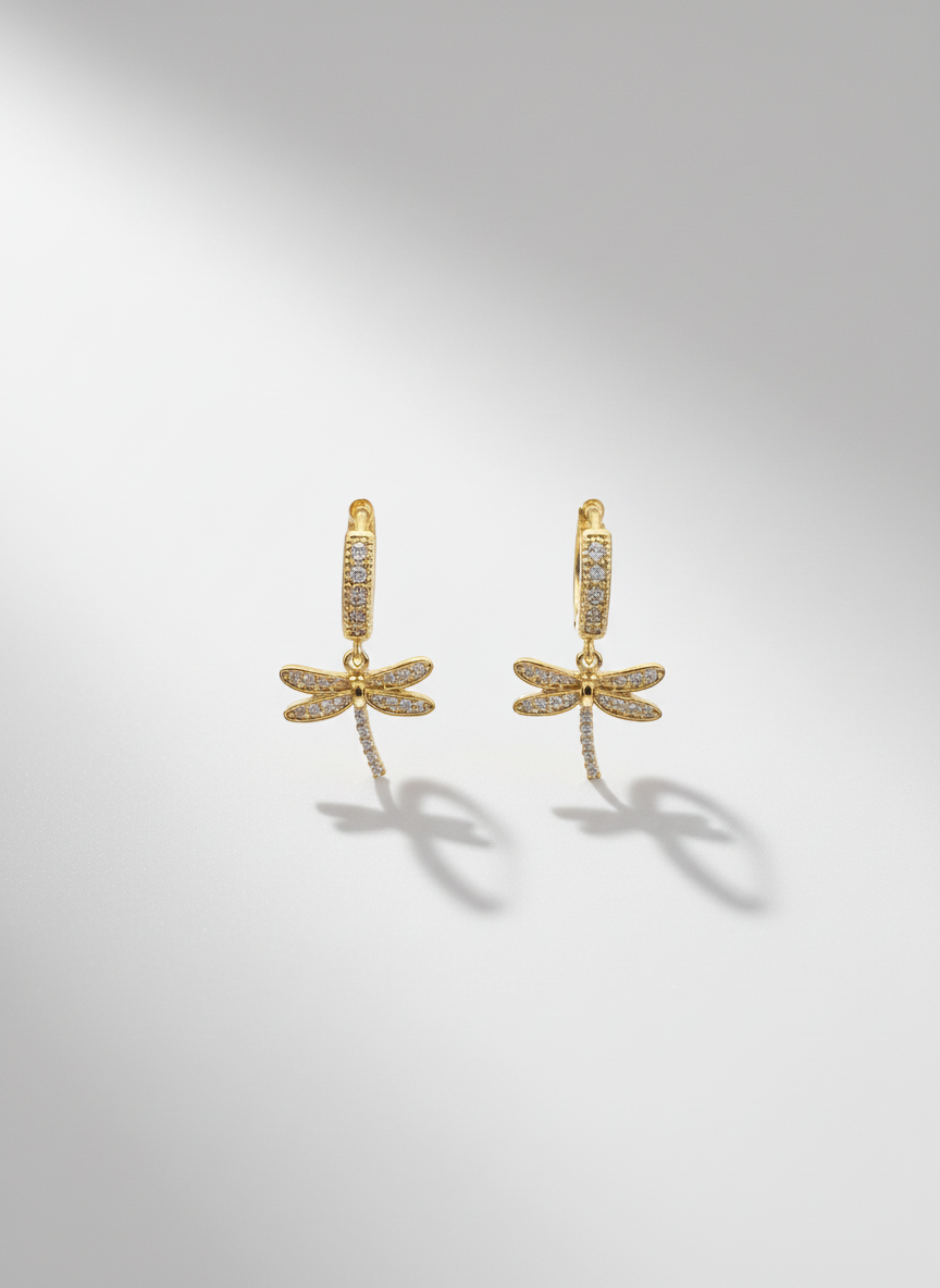 Close-up of Dragonfly Drops by Gracie Rose Creative, showcasing delicate gold huggies with sparkling dragonfly charms against a soft background.