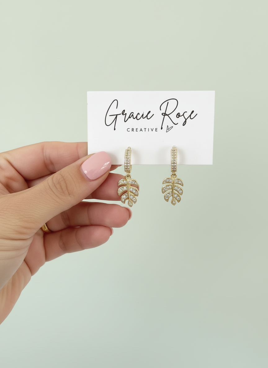 Stylish Monstera Shine Huggies by Gracie Rose Creative, showcasing sparkling crystal-studded leaf charms held by gold huggie hooks, elegantly displayed in a hand.