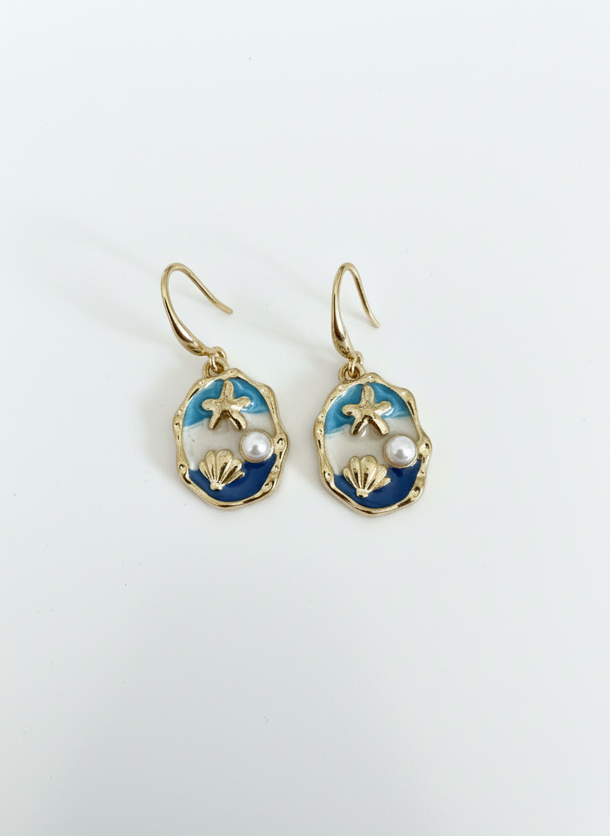 Handmade gold-toned earrings from Gracie Rose Creative called The Blue Horizon, featuring ocean-blue enamel, a pearl, starfish, and shell details.