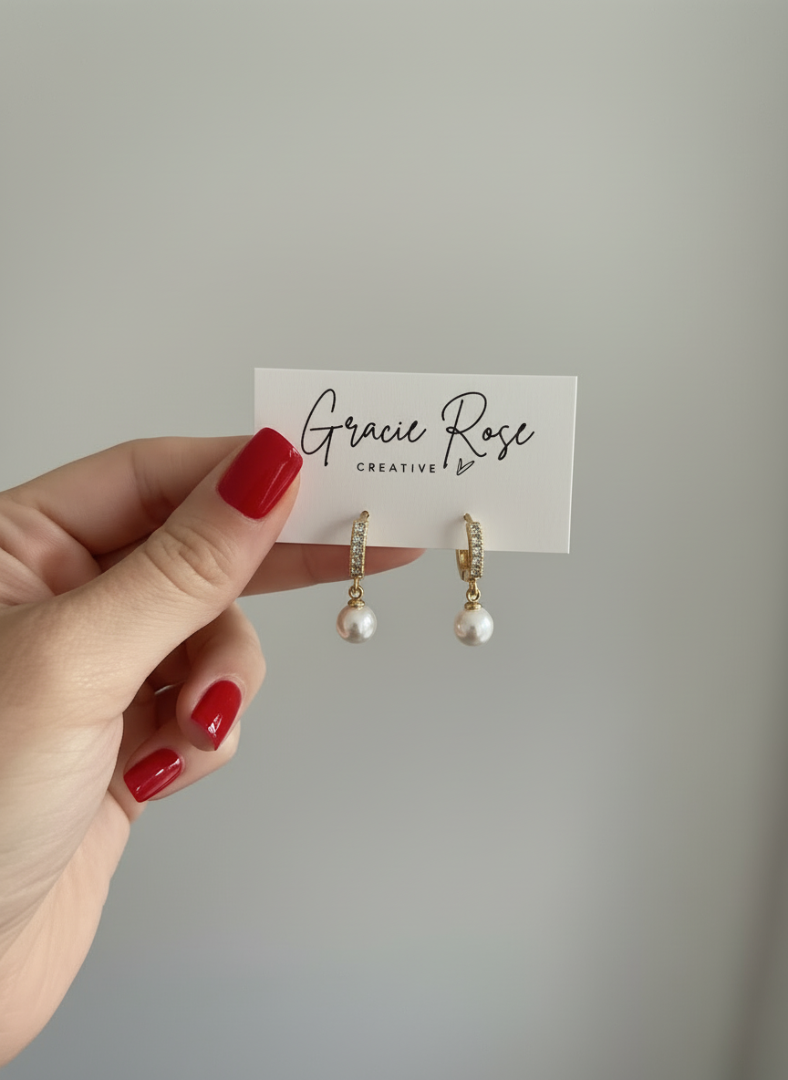 A hand holding a pair of Pearl Swing Huggies by Gracie Rose Creative, displayed on a branded card, emphasizing the unique pearl and gold details.