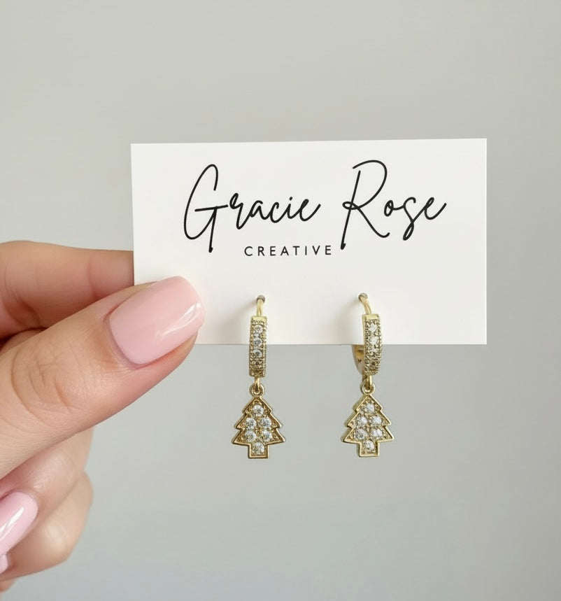 Hand holding Merry Little Trees earrings by Gracie Rose Creative, displaying the gold huggie hooks and charming Christmas tree design with glittering details.