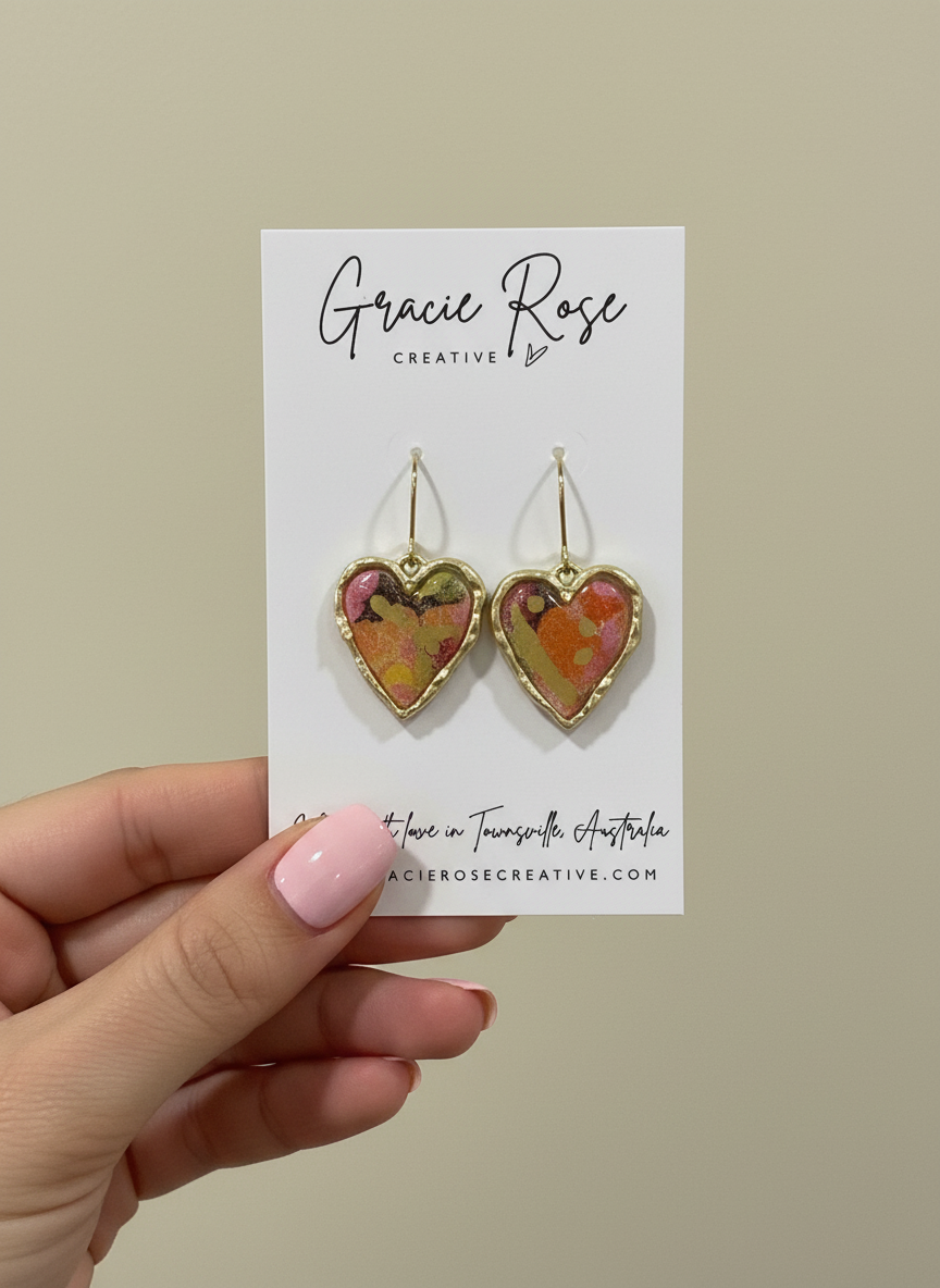 A hand holding a card displaying Rosé Hearts earrings by Gracie Rose Creative, featuring colorful heart-shaped polymer clay earrings with a unique design.