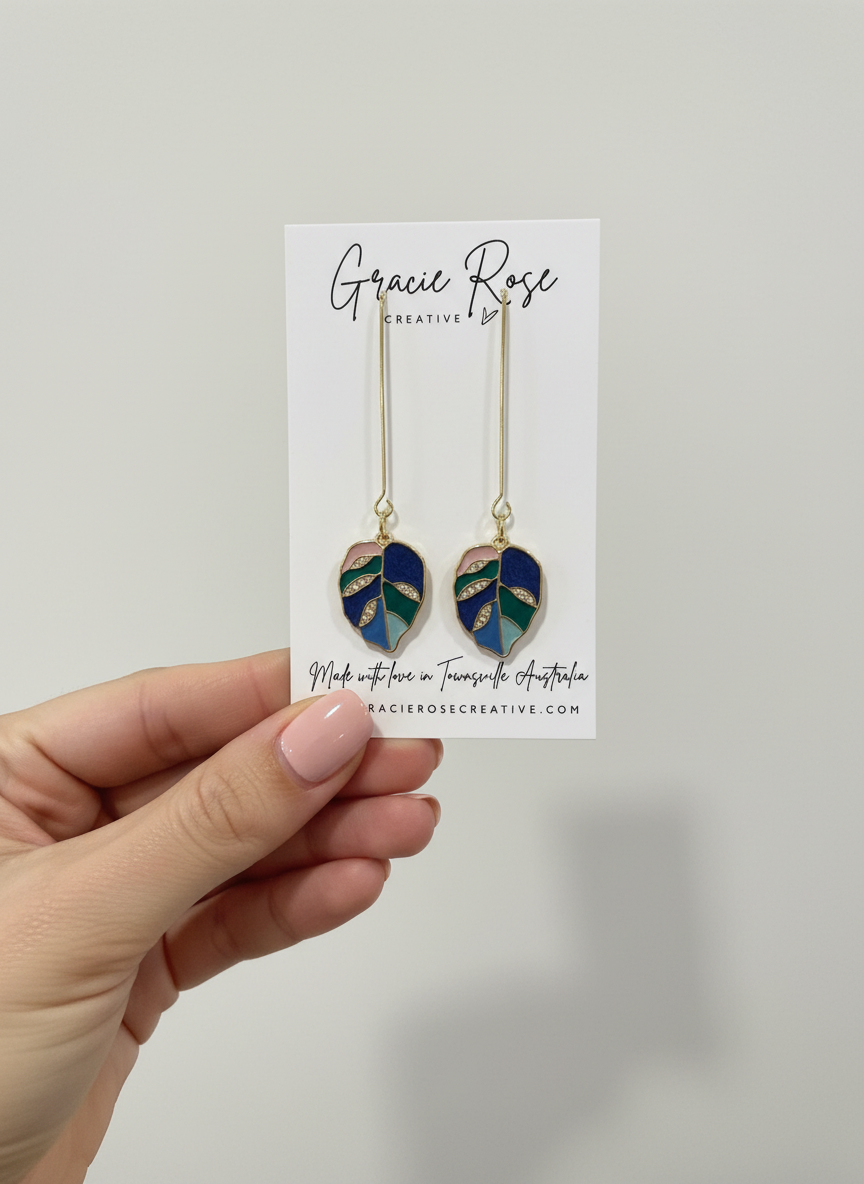 A hand holding Blue Bloom Leaves earrings on a branded card from Gracie Rose Creative. The earrings feature a unique leaf shape made with blue tones and gold accents, ideal for fashion lovers.