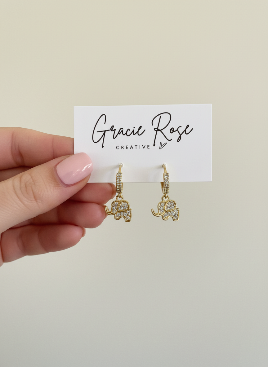 Hand holding gold huggie earrings with sparkling elephant charms, titled 'Lucky Elephant Drops' by Gracie Rose Creative.